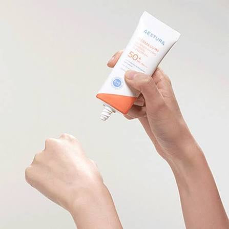 A hand holds a white and orange Aestura Sunscreen SPF 50+ tube above another hand, with sunscreen being squeezed onto the skin against a light gray background.