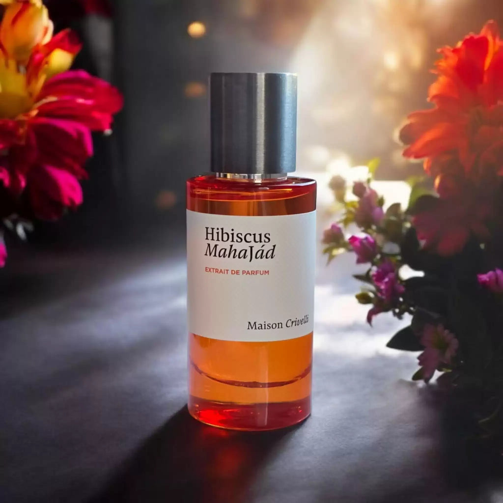 A bottle of Maison Crivelli Hibiscus Mahajad extrait de parfum with amber-colored liquid and a white label, set against a dark background with warm light flares and colorful flowers on both sides.