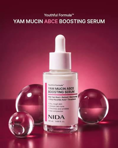 NIDA Yam Mucin Abce Boosting Serum in a clear glass dropper bottle with a white cap, set against a dark magenta background with three reflective glass spheres.