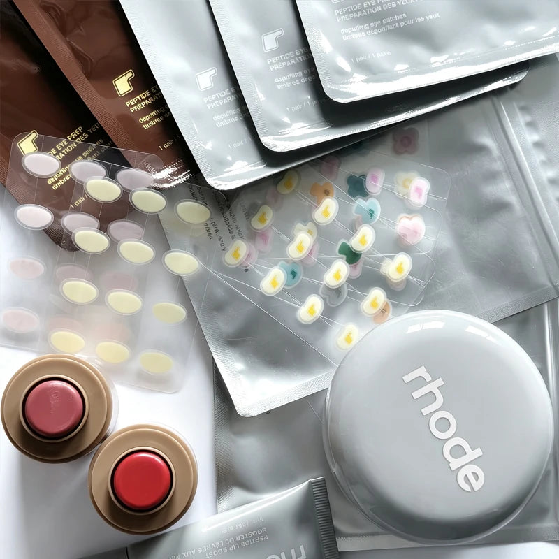 A styled flat-lay of skincare products, featuring a prominent silver round compact with the brand name 'rhode' printed in white, alongside two gold circular tubes with red and pink caps. Several silver and brown sachets, and sheets of small oval and heart-shaped patches are also arranged on a white surface.